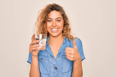 Genç, sarışın, mavi gözlü, beyaz arka planda bir bardak su içen, gülümseyen, işaret veren, parmaklarıyla başparmağını kaldıran güzel bir kadın.