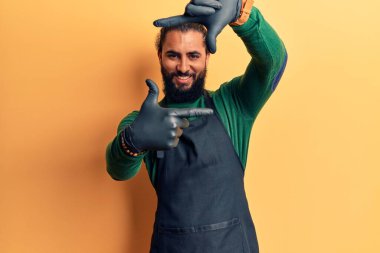 Genç Arap adam berber önlüğü takıyor gülümsüyor ve mutlu yüzlü eller ve parmaklarla çerçeve yapıyor. yaratıcılık ve fotoğrafçılık kavramı. 