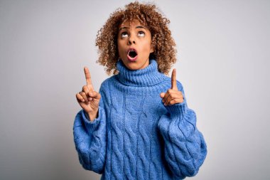 Beyaz arka plan üzerine balıkçı yaka kazak giyen güzel Afro-Amerikalı kadın hayret ve şaşkınlık içinde yukarı baktı ve parmaklarıyla işaret ve kollarını kaldırdı..