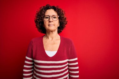 Orta yaşlı güzel kıvırcık saçlı kadın kırmızı arka planda sıradan çizgili süveter giyiyor yüzünde ciddi bir ifadeyle rahatlıyor. Basit ve doğal bir şekilde kameraya bakıyorsun..