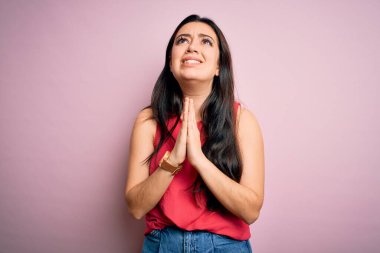 Genç esmer kadın pembe izole edilmiş arka plan üzerine gündelik bir tişört giyip elleri bağlı yalvarıyor ve dua ediyor. Yüzünde umut ifadesi var. Çok duygusal ve endişeli. Yalvarıyorum..