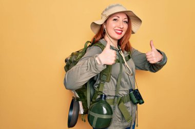 Sırt çantası ve sarı arka plan işareti üzerinde şapka takan kızıl saçlı genç bir kadın el hareketleriyle olumlu bir hareket yapıyor, gülümsüyor ve mutlu. Neşeli ifade ve kazanan jest.