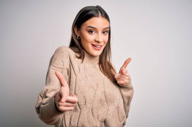 Genç, güzel esmer kadın, sıradan bir süveter giyip beyaz arka planda dikiliyor ve mutlu ve komik yüzüyle kamerayı işaret ediyor. İyi enerji ve titreşimler.