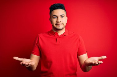 İzole edilmiş arka planda duran kırmızı polo giyen genç yakışıklı adam elleri ve kolları havada, kafası karışmış bir ifade takınıyor. Şüphe kavramı.