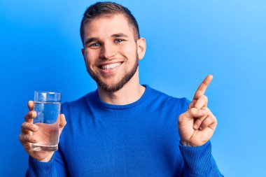 Genç yakışıklı adam, bir bardak su içerken gülümsüyor mutlu bir şekilde elini ve parmağını yana doğru gösteriyor. 