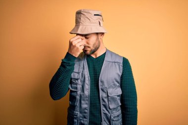 Tatilde sakallı, sarı arka planda kaşif şapkası takan yakışıklı turist yorgun burnu ve gözleri ovuşturup yorgunluk ve baş ağrısı çekiyor. Stres ve hüsran kavramı.