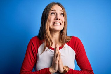 Genç, güzel, kızıl saçlı bir kadın, izole edilmiş mavi arka plan üzerine gündelik tişört giyip elleri bağlı yalvarıyor ve dua ediyor. Yüzünde umut ifadesi var. Çok duygusal ve endişeli. Yalvarıyorum..