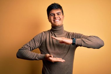 Sarıya boyanmış sarı arka plan üzerine sıradan kazak giyen genç yakışıklı bir adam. Elleri büyük ve büyük boy işareti, ölçü sembolü gösteriyor. Kameraya bakıp gülümsüyor. Ölçüm konsepti.