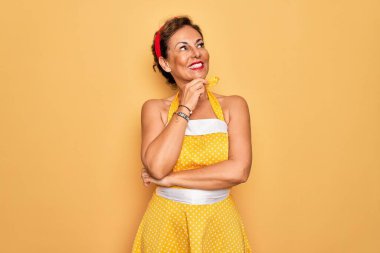 50 'li yaşlarda, sarı arka plan üzerine eski tarz elbise giyen, elleri çenesinde soru soran, düşünceli ifadeli orta yaşlı bir kadın. Gülen ve düşünceli bir yüz. Şüphe kavramı.