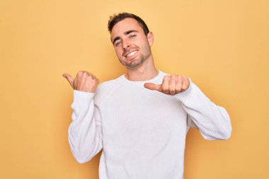 Mavi gözlü, sıradan bir süveter giyen, sarı arka planda duran, el ve baş parmakları havada, kendine güvenen genç, yakışıklı bir adam.