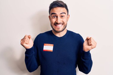 Sakallı yakışıklı genç adam, izole edilmiş beyaz arka planda kimlik etiketi takıyor. Gururla bağırıyor, zaferi kutluyor ve kollarını kaldırarak başarıyı kutluyor.