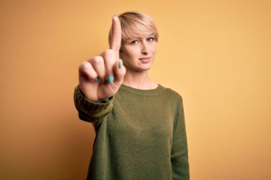 Modern kısa saçlı, sarı arka planda sıradan bir süveter giyen genç, sarışın bir kadın. Parmağını kaldırmış ve kızgın bir ifadeyle işaret ediyor.