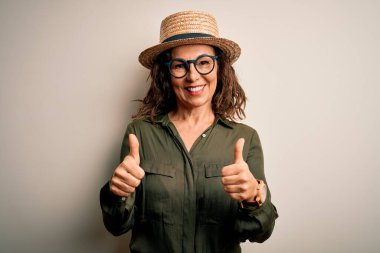 Orta yaşlı esmer kadın gözlüklü ve şapkalı, izole edilmiş beyaz arka plan burcunun üzerinde duruyor. Elleriyle olumlu jest yapıyor, gülümsüyor ve mutlu. Neşeli ifade ve kazanan jest.