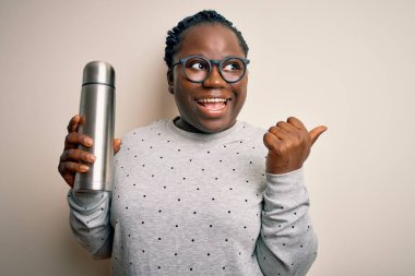 Genç Afro-Amerikalı, örgülü, termosu olan, elinde kahve tutan ve gülümseyen yüzüyle baş parmağıyla yüzünü gösteren iri yarı bir kadın.