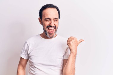 Orta yaşlı yakışıklı bir adam, günlük tişört giyip izole edilmiş beyaz arka planda dikiliyor başparmağıyla yan tarafa bakıyor ve ağzı açık gülümsüyor.