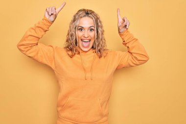 Sarışın, güzel, sportif bir kadın sarı arka planda rahat bir tişört giyiyor. Şaşırmış, şaşırmış ve parmaklarıyla işaret ediyor ve kollarını kaldırıyor..