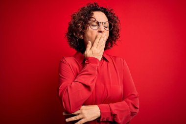 Orta yaşlı güzel kıvırcık saçlı kadın kırmızı arka planda günlük gömlek ve gözlük takıyor. Elle ağzını kapatmaktan sıkılmış. Huzursuz ve uykulu..