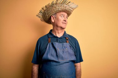 Orta yaşlı, beyaz önlüklü ve şapkalı bir adam izole edilmiş sarı arka planda, yüzünde gülümseme, doğal bir ifade ile yan yana bakıyor. Kendinden emin gülen.