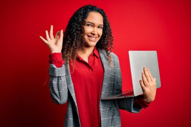 Kıvırcık saçlı genç bir iş kadını kırmızı arka planda dizüstü bilgisayar kullanıyor. Tamam işareti yapıyor, mükemmel bir sembol.