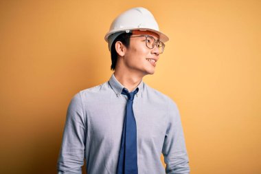 Güvenlik kaskı takan ve sarı arka planda kravat takan genç yakışıklı mimar yüzünde gülümseme, doğal bir ifade ile diğer tarafa bakıyor. Kendinden emin gülen.