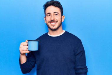Genç İspanyol adam elinde kahve tutarken pozitif ve mutlu görünüyor. Durup gülümsüyor ve kendine güvenen bir gülümsemeyle dişlerini gösteriyor. 