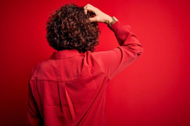 Orta yaşlı güzel kıvırcık saçlı kadın kırmızı arka planda günlük gömlek ve gözlük takıyor. Geriye doğru, elleri kafasında şüpheyi düşünüyor.