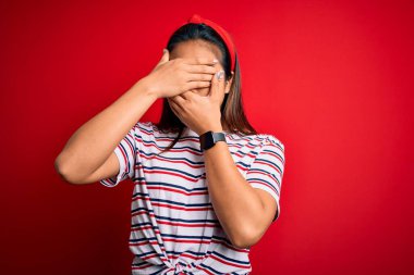 Genç, güzel Asyalı bir kız izole edilmiş kırmızı arka plan üzerinde sıradan çizgili tişört giyiyor gözleri ve ağzı kapalı, şaşırmış ve şok olmuş. Duygularını gizliyor.