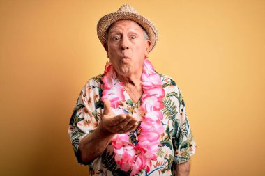 Beyaz saçlı, yaz şapkası takan ve sarı arka planda Hawaii zambağı takmış kameraya bakıp havada öpücükler savuran hoş ve seksi bir adam. Aşk ifadesi.