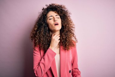 Kıvırcık saçlı ve piercingli genç bir iş kadını. Zarif bir ceket giyiyor. Acılı boynuna dokunuyor.