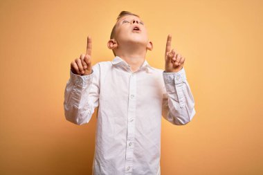 Küçük, beyaz, mavi gözlü, sarı arka planda zarif beyaz gömlek giyen genç bir çocuk. Şaşırmış ve şaşırmış bir şekilde yukarı bakıyor, parmaklarıyla işaret ediyor ve kollarını kaldırıyordu..