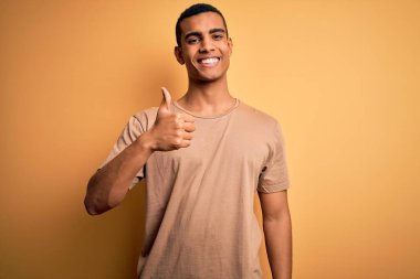 Sarı arka plan üzerinde sıradan tişörtler giyen genç ve yakışıklı Afro-Amerikan bir adam elleriyle mutlu bir jest yapıyor. Kameraya bakıp başarıyı gösteren ifade onaylanıyor.