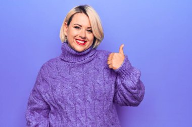 Güzel sarışın ve cüsseli bir kadın mor arka planda balıkçı yaka kazak giyip mutlu bir el hareketi yapıyor. Kameraya bakıp başarıyı gösteren ifade onaylanıyor.