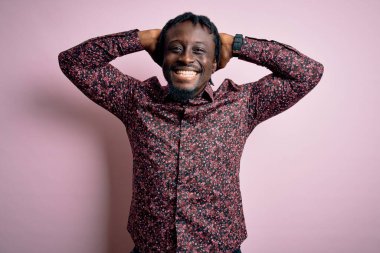 Genç, yakışıklı, Afro-Amerikalı bir adam pembe arka planda dikilmiş kolları ve elleri başının ve boynunun arkasında mutlu bir şekilde gülümsüyor.