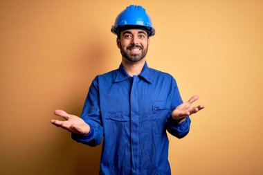 Sakallı tamirci mavi üniformalı ve kasklı sarı arka planda gülümseyen neşeli kolları açık hoş geldin, olumlu ve kendinden emin selamlar.