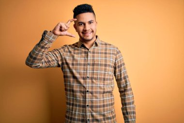 İzole edilmiş sarı arka plan üzerinde duran sıradan gömlekli genç yakışıklı adam gülümsüyor ve el hareketleriyle küçük beden işareti yaparak kameraya bakıyor. Ölçü kavramı.