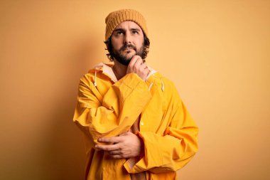 Yağmurlu bir günde yağmurluk giyen sakallı genç yakışıklı bir adam sarı arka planda elleri çenesinde soru sorarken düşünceli bir ifade takınıyor. Düşünceli bir yüzle gülümsüyor. Şüphe kavramı.