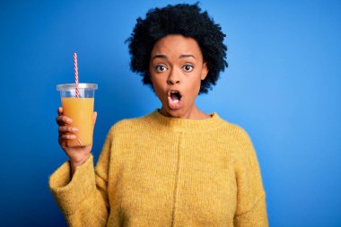 Kıvırcık saçlı, sağlıklı portakal suyu içen genç Afrikalı Afro kadın şaşırmış bir yüzle korkmuş ve korku ifadesinden heyecanlanmış.