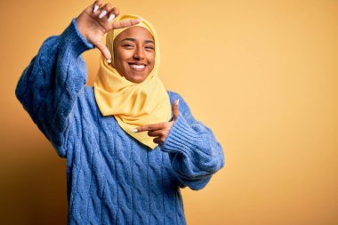 Genç Afrikalı Afro kadın, izole edilmiş sarı arka planda Müslüman başörtüsü takıyor gülümsüyor ve mutlu yüzlü elleri ve parmakları olan bir çerçeve yapıyor. Yaratıcılık ve fotoğrafçılık kavramı.