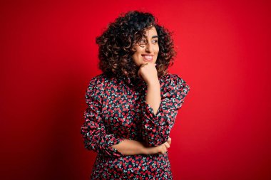 Genç, güzel, kıvırcık, çiçekli bir elbise giyen, elleri çenesinde kırmızı arka plan üzerinde duran, soru soran, düşünceli bir kadın. Düşünceli bir yüzle gülümsüyor. Şüphe kavramı.