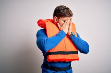 Sakallı ve mavi gözlü genç bir turist beyaz arka planda can yeleği giyiyor ve ağlarken suratını ellerle kapatıyor. Depresyon kavramı.