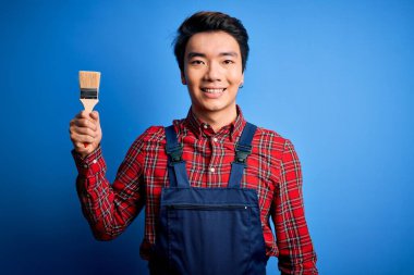 Önlük giyen genç, yakışıklı Çinli ressam mutlu yüzlü fırça kullanıyor. Dişlerini gösteren bir gülümsemeyle ayakta duruyor.