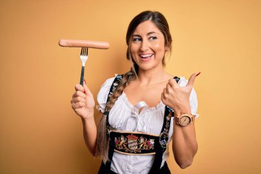 Genç esmer Alman kadın geleneksel Ekim Festivali elbisesi giyiyor. Elinde sosis var ve gülümseyen bir gülümsemeyle baş parmağıyla onu gösteriyor.