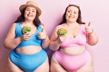 Bikini giyen, elinde brokoli olan, mutlu ve pozitif gülümseyen, başparmağıyla mükemmel ve onay işareti yapan genç ve büyük beden ikizler. 