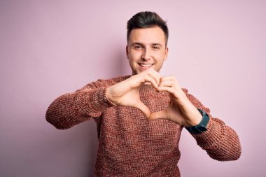 Pembe izole edilmiş arka plan üzerine sıradan bir kazak giyen yakışıklı beyaz bir adam elleriyle kalp sembolü yapan bir aşkla gülümsüyor. Romantik konsept.