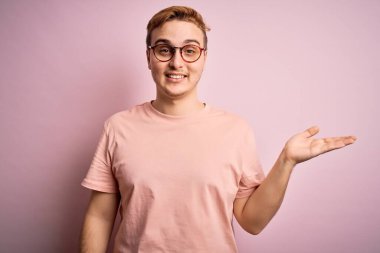 Genç, yakışıklı, günlük tişört giyen, pembe arka plan üzerinde duran neşeli bir şekilde gülümseyen ve avuç içi kameraya bakan bir adam..