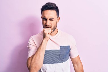 Sakallı, sıradan tişörtlü, pembe arka planda duran, kendini kötü hisseden ve soğuk algınlığı veya bronşitin belirtisi olarak öksüren genç yakışıklı bir adam. Sağlık hizmeti kavramı.