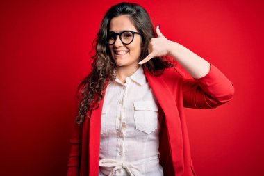 Kıvırcık saçlı, ceketli, kırmızı arka planda gözlüklü, gülümseyen, el ve parmaklarıyla telefonla konuşan güzel bir kadın. İletişim kavramları.