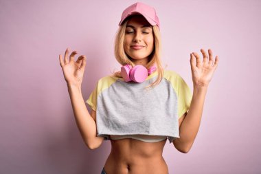 Genç ve güzel sarışın sporcu. Pembe arka planda kulaklık takarak spor yapıyor. Gözleri kapalı gülümsüyor ve parmaklarıyla meditasyon yapıyor. Yoga konsepti.