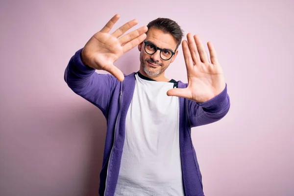 手の手のひらと指を使用してフレームを行うピンクの背景に立って紫のスウェットシャツと眼鏡を身に着けている若いハンサムな男 カメラの視点 — ストック写真