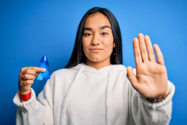 年轻的亚洲女人拿着蓝色的癌症彩带标志站在孤立的背景上 张开手做停止手势 严肃而自信地表达着防守姿态 — 图库照片